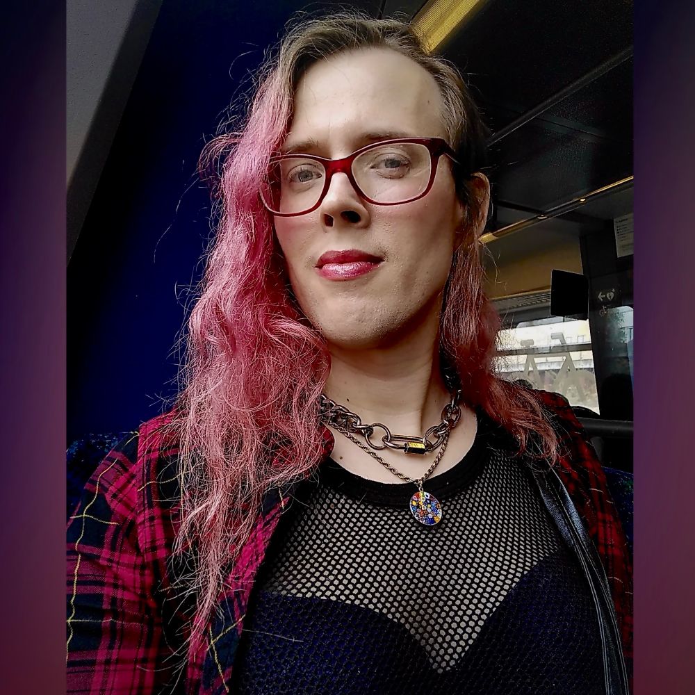 
This is a selfie I took in a the train, you can only see the top part of me. I’m smiling and looking down in a slight angle towards the camera that I’m also holding a bit down. I have long light red hair, mostly swept to one side. I’m wearing red glasses. I have makeup on with a subtle blush on my cheeks and a red lipstick with a shiny silver glass on top. 

I’m wearing a red checkered shirt that is open in the front and I have a net blouse underneath, and you can see my purple bra I’m wearing through the net blouse. I’m wearing a heavy chain necklace that is short, almost like a choker, and a silver necklace with a pendant from Italy, its a metal band with some assorted colors of glass melted into it. 

In the background, you can see some of the train interior, but it’s a bit uncertain what is what, unless you specifically know how Danish train interior looks like.