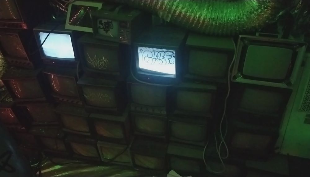 a stack of old monitors marked with grafitti. two are on. a ventilation pipe runs above. 