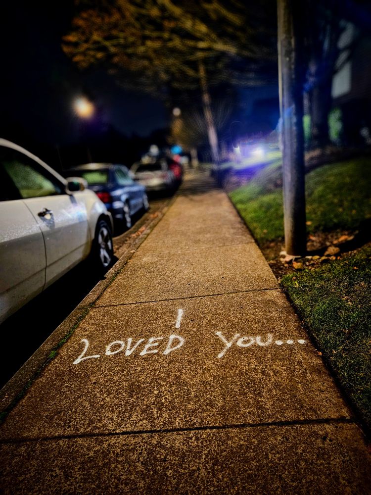 A picture of the phrase "I loved you ..." written in white chalk on the sidewalk at night. 