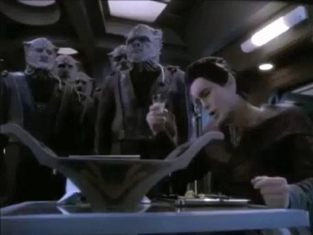 a group of aliens are gathered around a table with a woman reading a book .