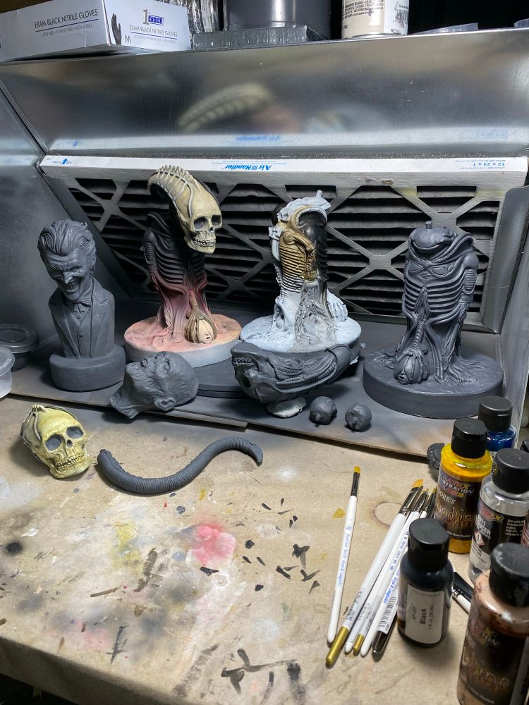 A series of maquette’s and maquette pieces in a desktop spray booth. In the foreground are some paintbrushes and bottles of paint. The tabletop is covered in paint spills and brush strokes in various colors. Some of the maquette’s are partially painted: some with dark blacks and flesh tones, others in bone color and black washes. Some are primed in dark gray primer. The maquette’s range from a laughing clown reminiscent of Joker, a skull, a biomechanical, and a zombie.