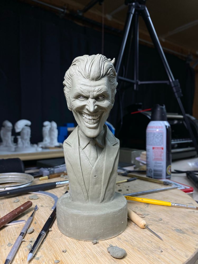 A clay representation of the comic and movie character “The Joker”. Made in Chavant NSP clay. A menacing Joker glares at the viewer with  a cheek to cheek smile. He wears a suit, and his hair is swept back. The bust of this figure connects to a circular base made of the same clay. 