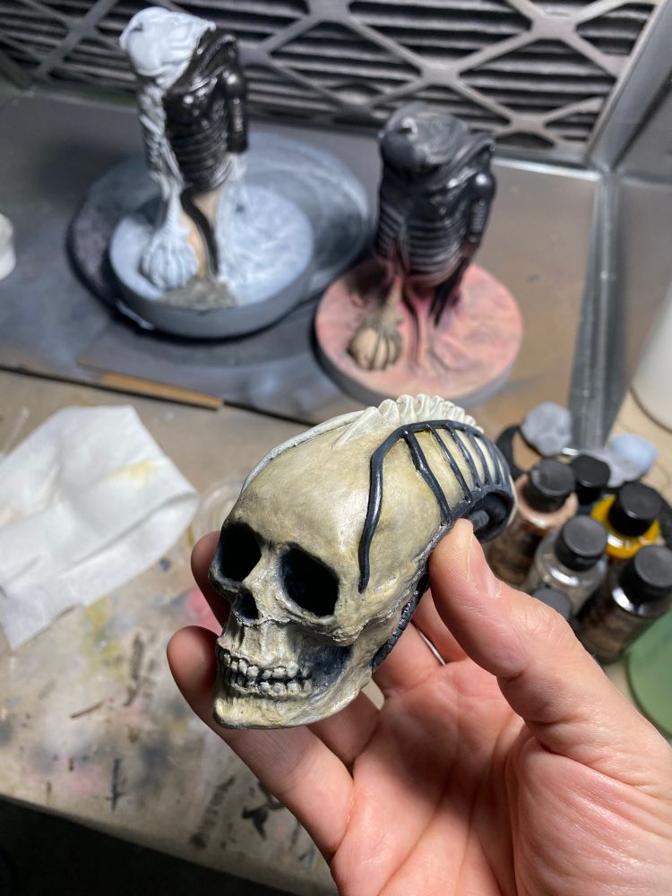 Biomechanical skull head held up to the camera. Painted a bone color with black accents. I’m the background are bottles of paint and two biomechanical bodies with test paint applications. 