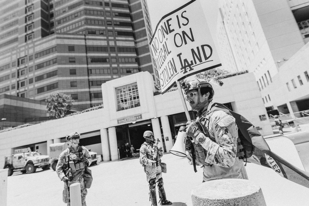 A protester wearing a Marine MARPAT uniform walks in front of Marines with a sign that reads “NO ONE IS ILLEGAL ON STOLEN LAND” (BW)