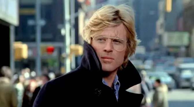 Robert Redford in Three Days of the Condor. A blonde white man with large glasses wearing a dark blue overcoat. He is standing on a busy street.