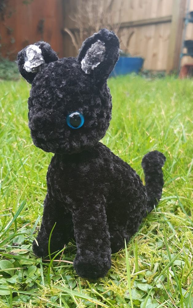 Black crocheted kitten sitting in grass, facing towards the camera. He has blue eyes & grey inside his ears.