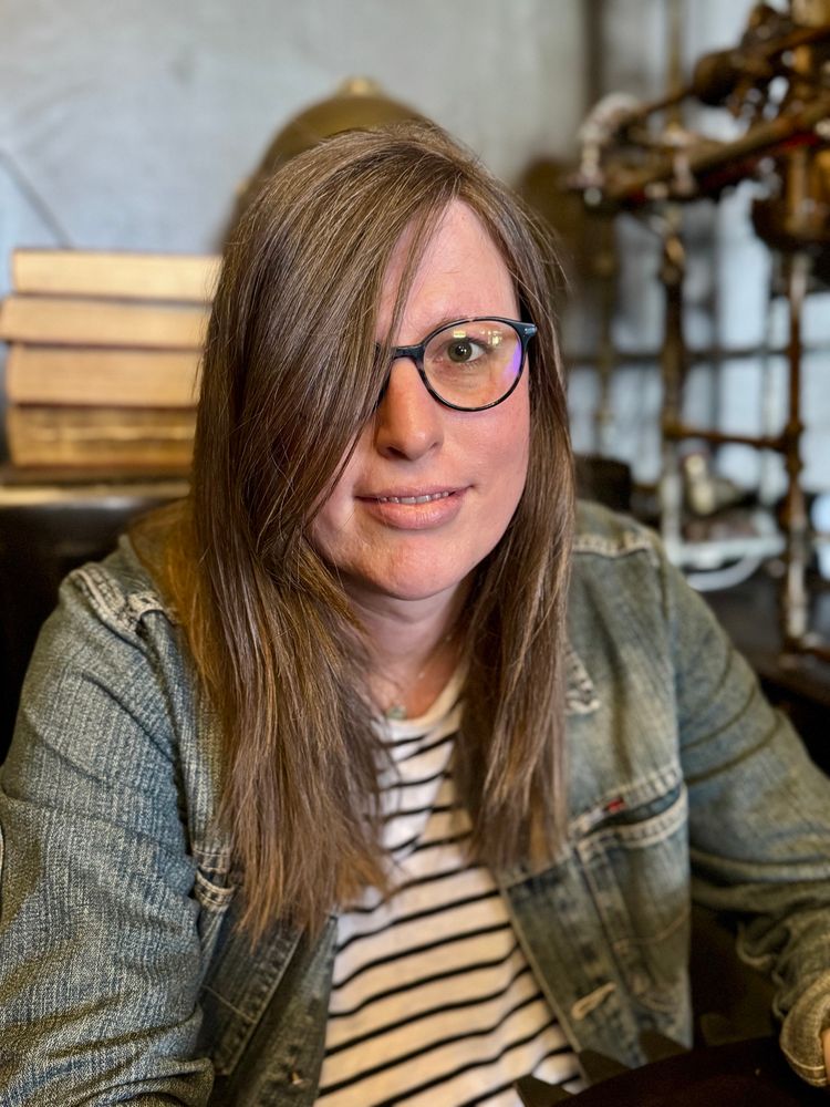 woman with glasses, looking at the camera, she is wearing a denim jacket and a stripe tshirt, she has long hair, covering one part of her face