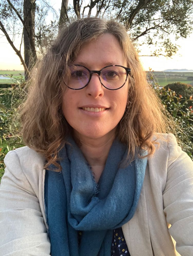 a selfie of a queer trans girl; the girl is wearing a beige jacket with a blue flower blouse and a sea blue scarf. She is also wearing big glasses.