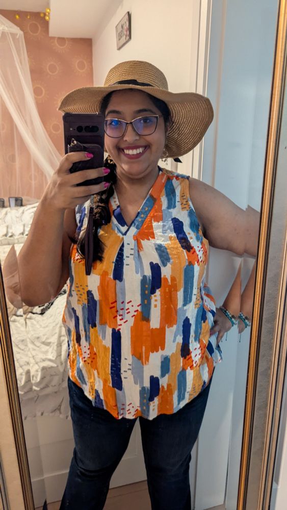 Full mirror selfie of Leona with dark jeans, a sleeveless blouse with a blue, white and orange pattern, her hair in a braid, makeup and earrings and a big sun hat.