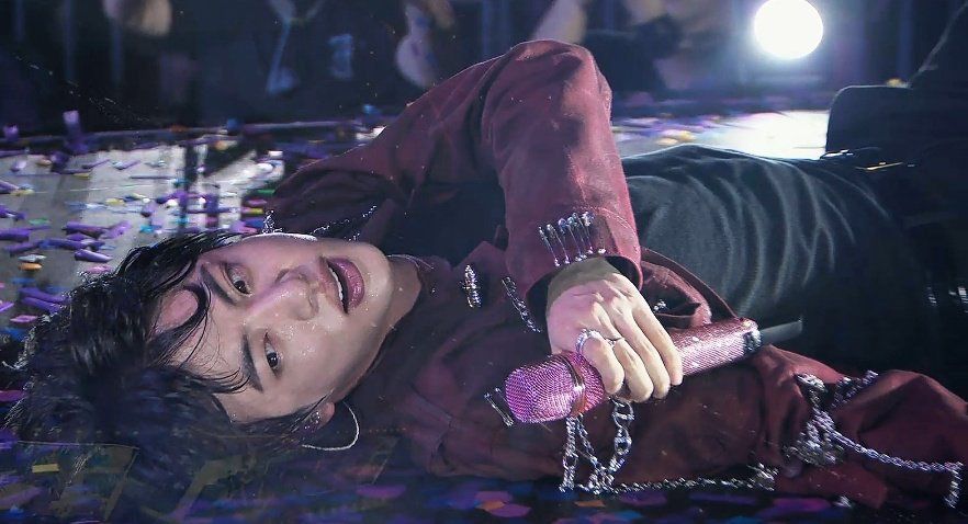 screenshot of Jin lying on the floor while singing Astronaut.. he is wearing a black tank top and burgundy jacket and looks absolutely otherworldly beautiful 
