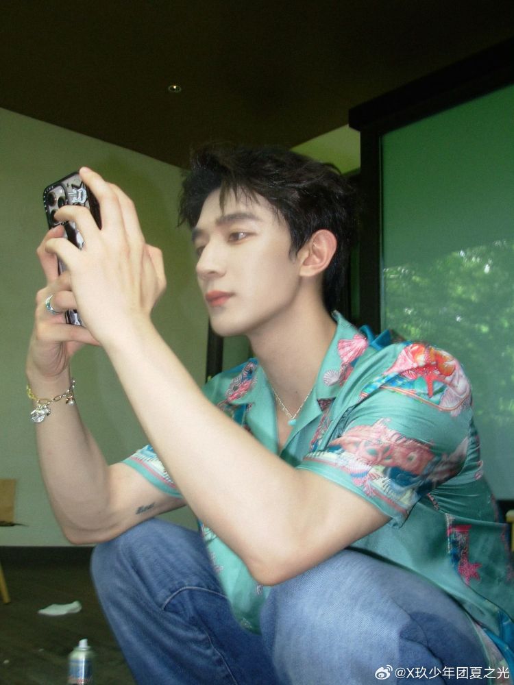 xia zhiguang in a silk teal shirt with pink and blue seashell designs knelling with his elbows resting on his knees looking at his cellphone. 