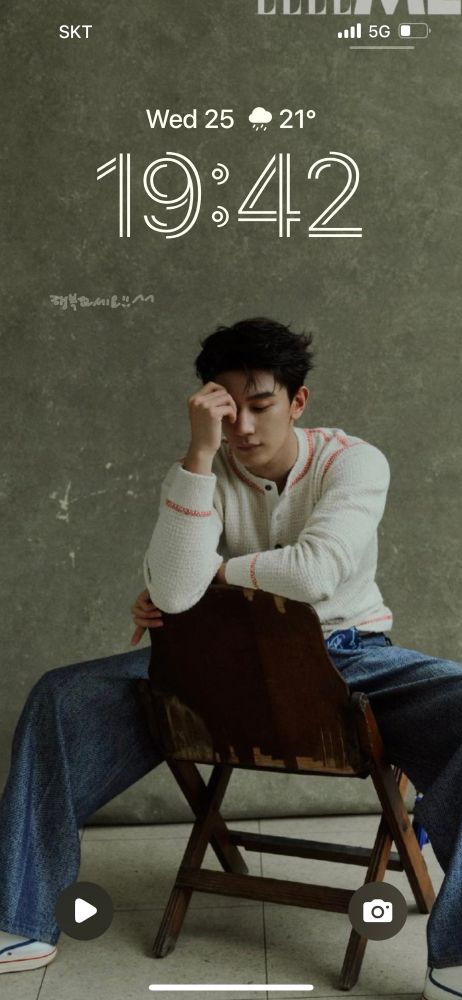 screenshot of a phone lock screen with xia zhiguang sitting on a backwards wooden chair in a white knit shirt and jeans. 