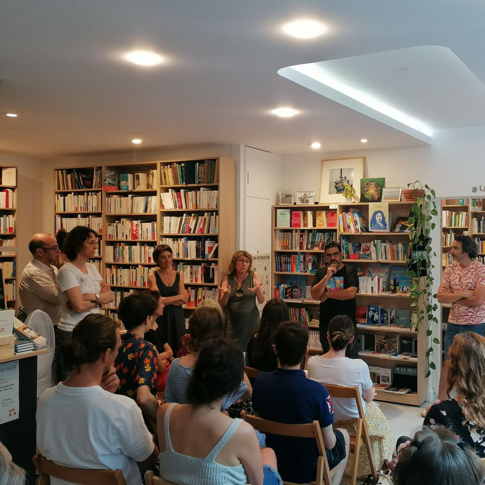 Photo carrée de l'intérieur d'une librairie, sept personnes sont debout et font face à des personnes assises sur des chaises en bois. 