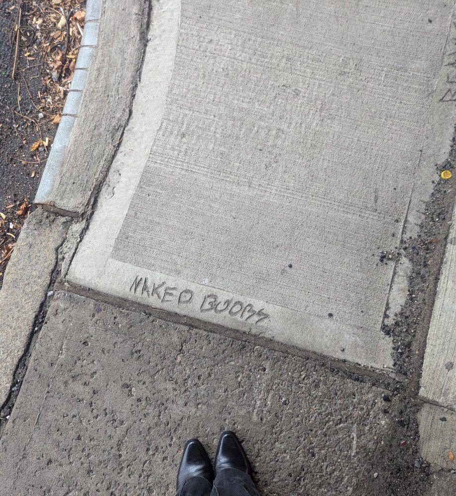 Photo of some concrete that someone has written "naked boobs" in. Also the tips of my boots.