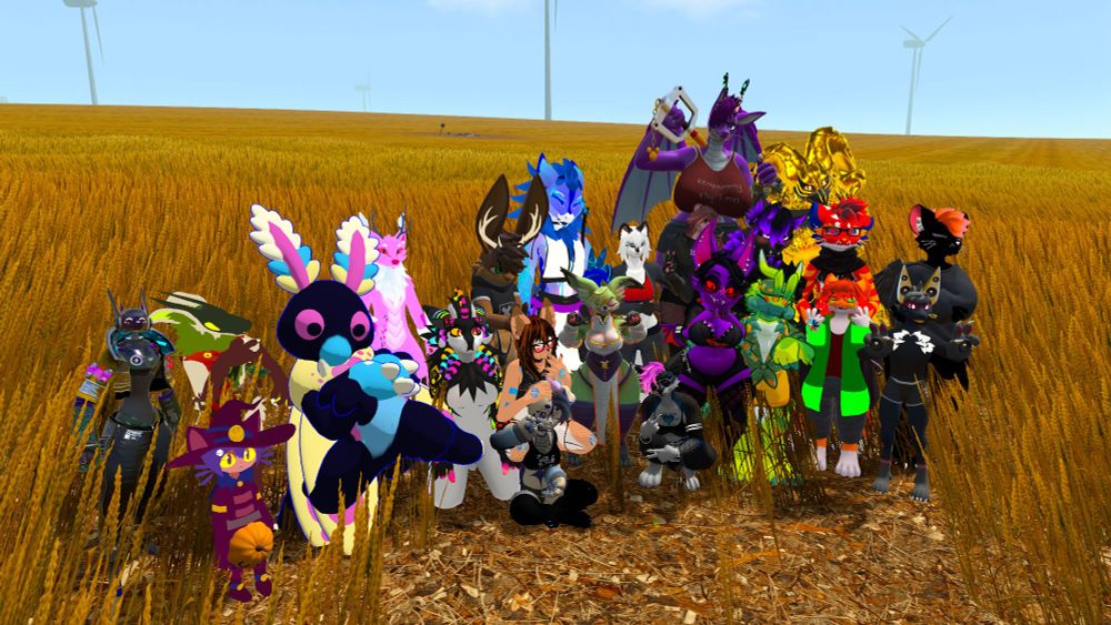 Group picture in VRChat. The setting is a seemingly endless expanse of wheat, with some wind turbines in the background. In the foreground are a dozen or so very colorful anthropomorphic animal avatars.