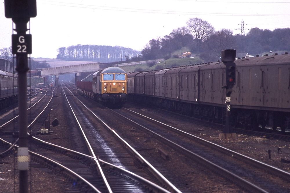 A scene at Grantham station, April 1977. Tracks in foreground, a signal at the left hand side, coaches at the right, a train approaching and trees in the distance.