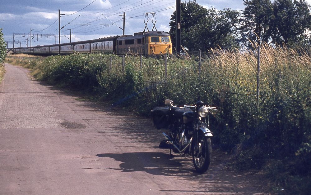 A road heads off into the left hand distance. A Vincent motorbike in the foreground and a train approaching.