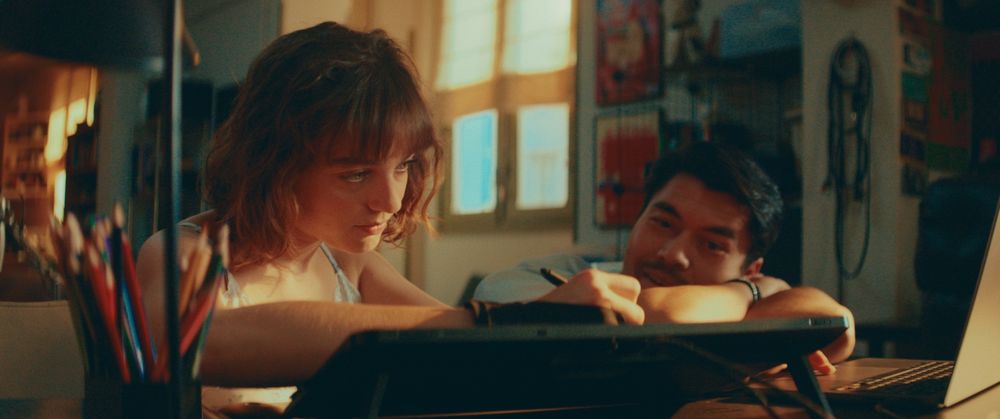 Fotograma de la película en el que se ve, de izquierda a derecha, a Beatrice Grannò, dibujando con un lápiz electrónico y mirando a la pantalla de un ordenador portátil, y a Henry Golding, observando cómo trabaja.