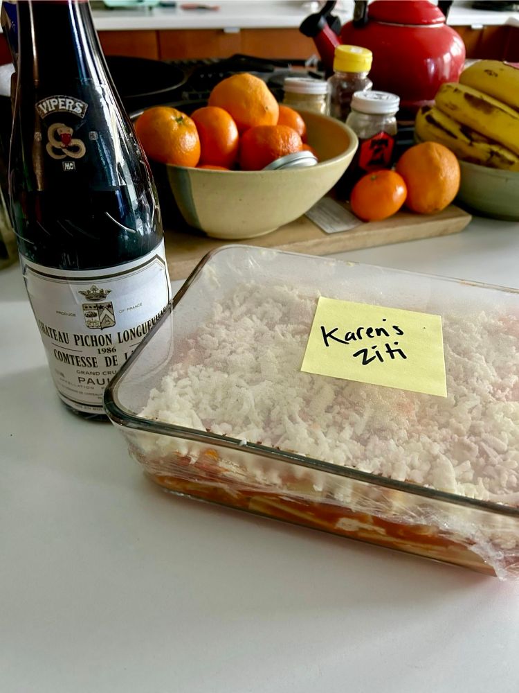 A tray of baked ziti with post it reading “Karen’s Ziti” and a bottle of wine w a taped on label for the “1986 Chateau Pichon Longueville Comtesse de Lalande” and a “Vipers” motorcycle gang insignia. 