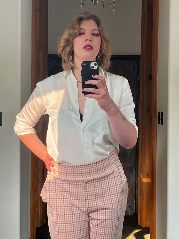 Another photo of Zefyr, a white trans femme wearing plaid pants and a silk shirt, posing in the mirror of an opulent bathroom 