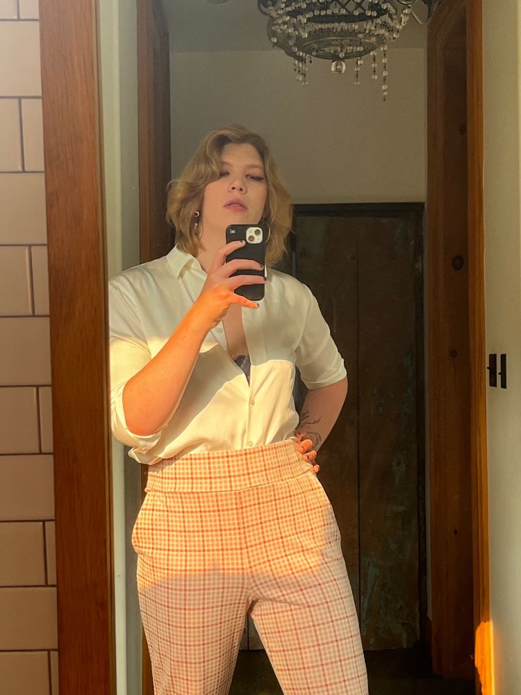 Zefyr, a white trans femme wearing plaid pants and a silk shirt, posing in the mirror of an opulent bathroom 