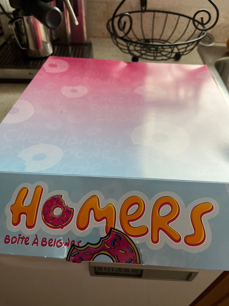 A closed box that is pale blue and pink with the writing “HOMERS boîte à beignes » written in pink and yellow letters in the Simpsons font. 
