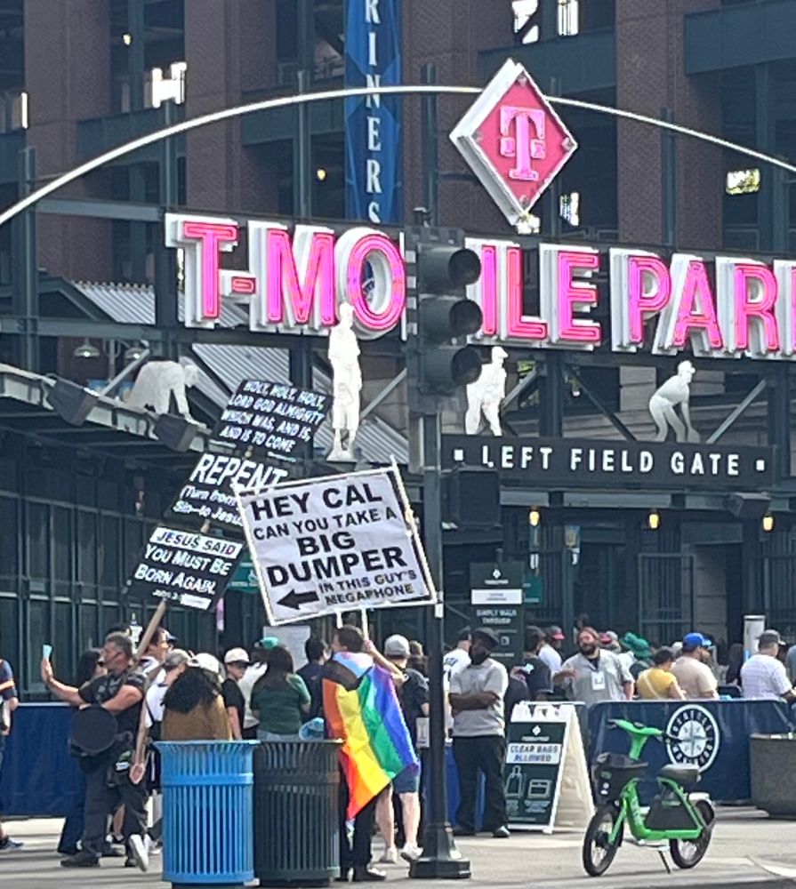 Outside T-Mobile Park. Awesome dude standing next to the terrible Jesus megaphone man, wearing a Pride flag and carrying a sign that says, “hey Cal can you take a big dumper in this guy’s megaphone”. 