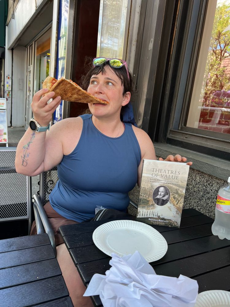 A woman in a blue tank top and brown skirt sits at a table, eating a slice of pizza. She’s holding a copy of a book, Theatres of Value. 