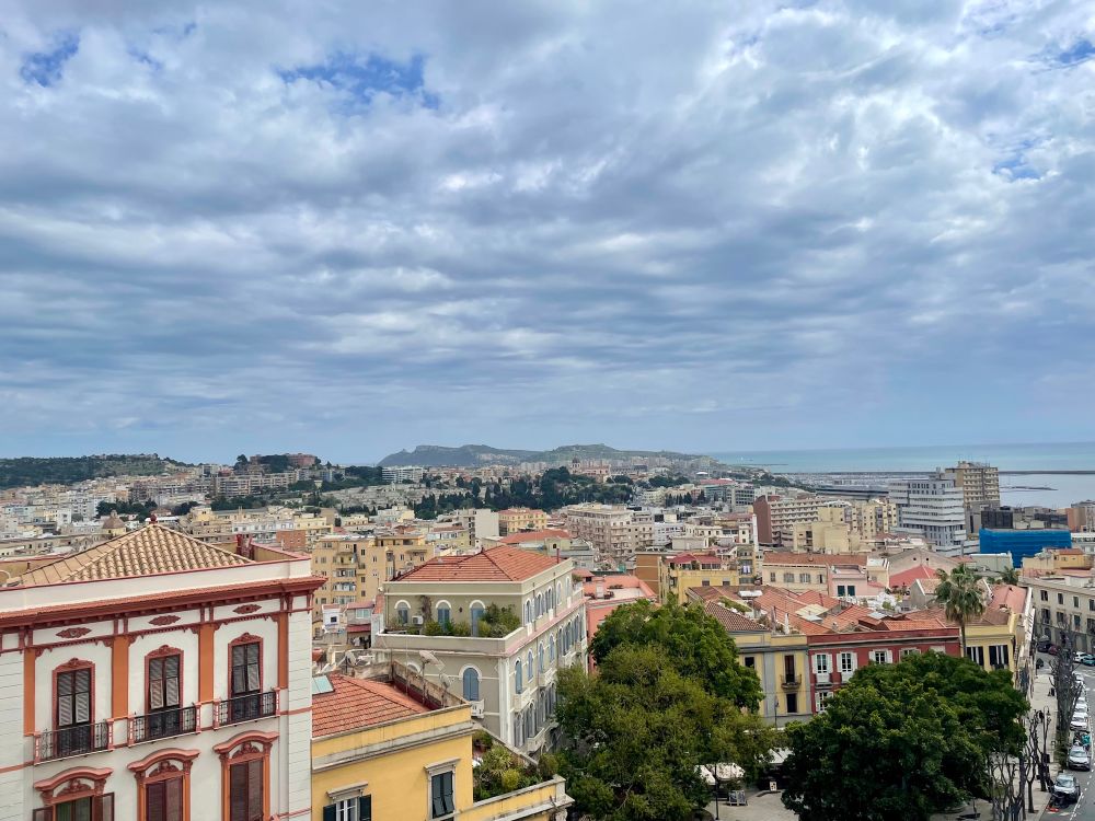 A view of the city of Cagliari, showing varying levels of buildings, a cloudy sky, and the ocean.