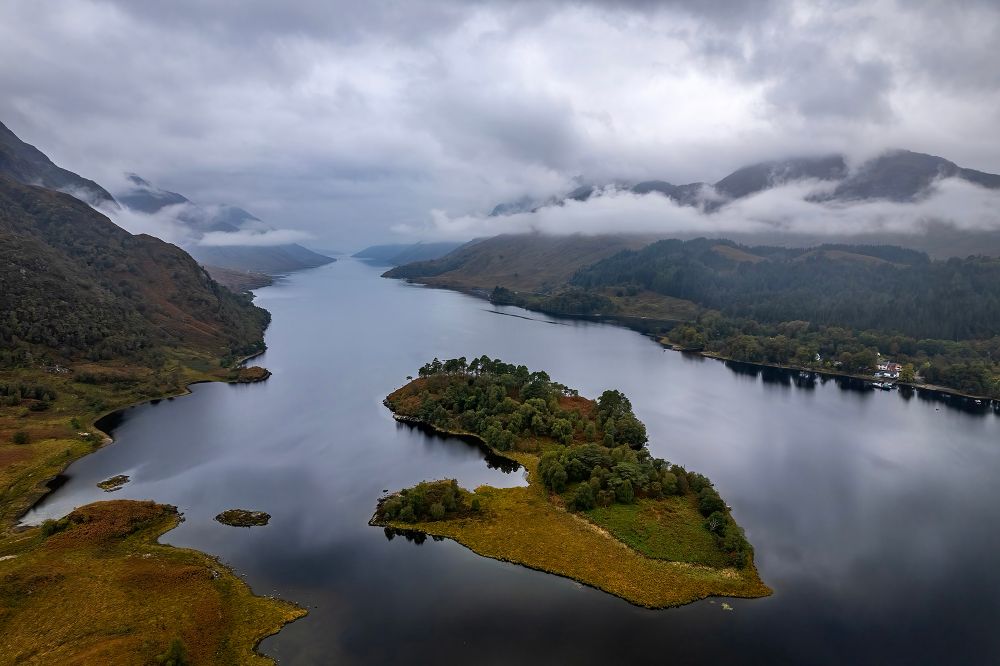 A view down a Scottish loch shows a tree-covered island in the foreground. At the right are steep hills descending to the water and low clouds partially obscure that hills at the right side. 