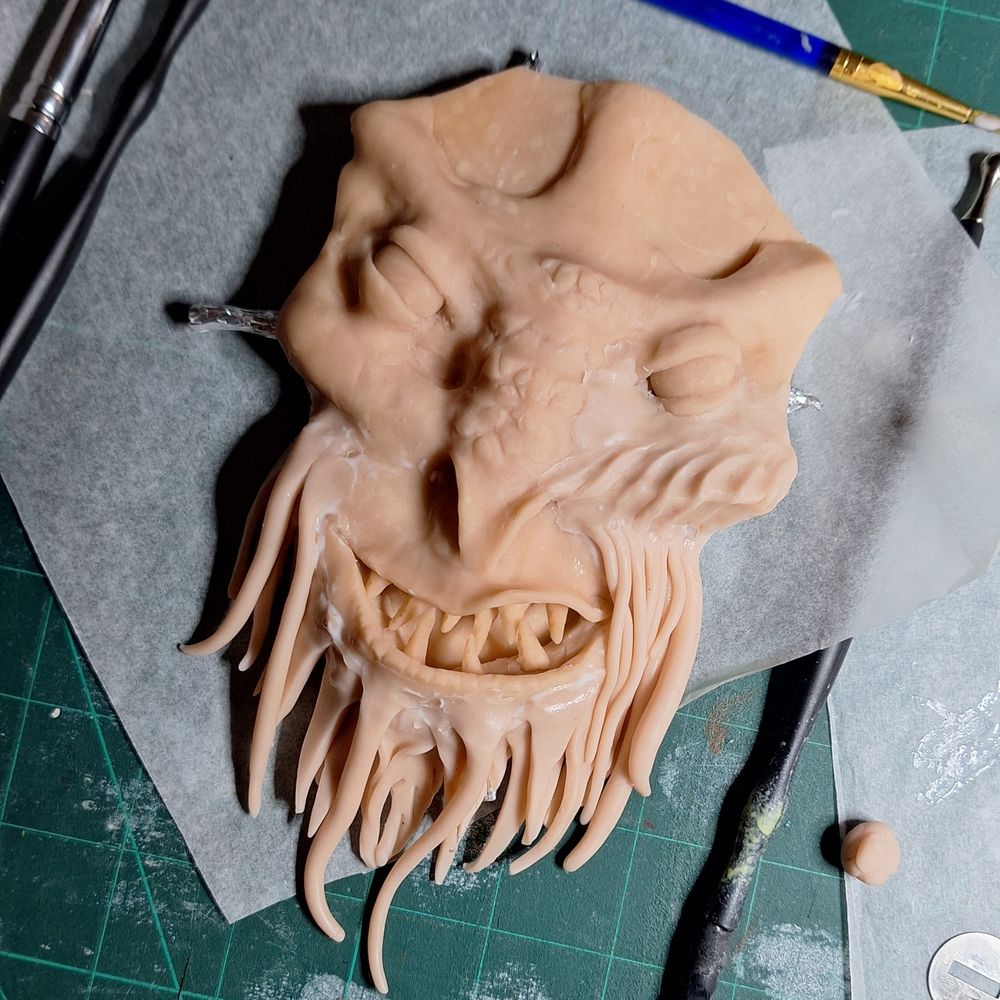 A face in pink sculpy. It's grimacing, has pointed teeth, scaley nose, and ridged cheek bones. It is unfinished.