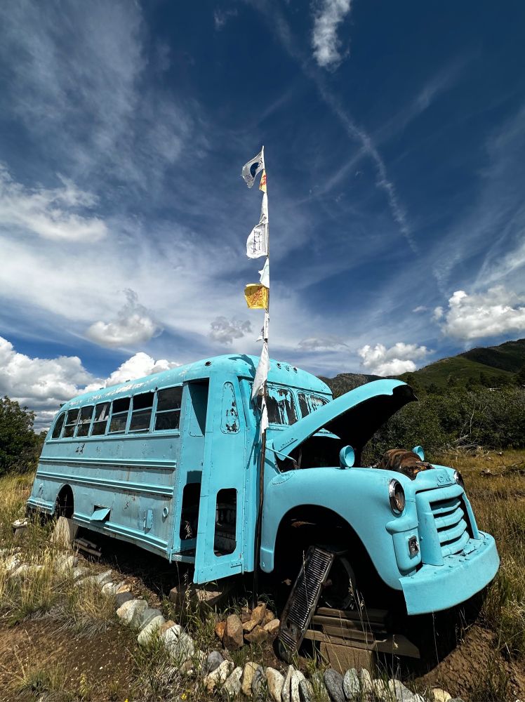 A broken down blue bus with a prayer flag pole in a field in hilly terrain.