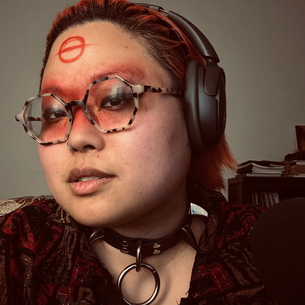 Josephine selfie with a gritty and dark filter: red eyeshadow messily all over her eyes with an alchemical symbol across her forehead. She’s also wearing a spiked collar with a silver ring.