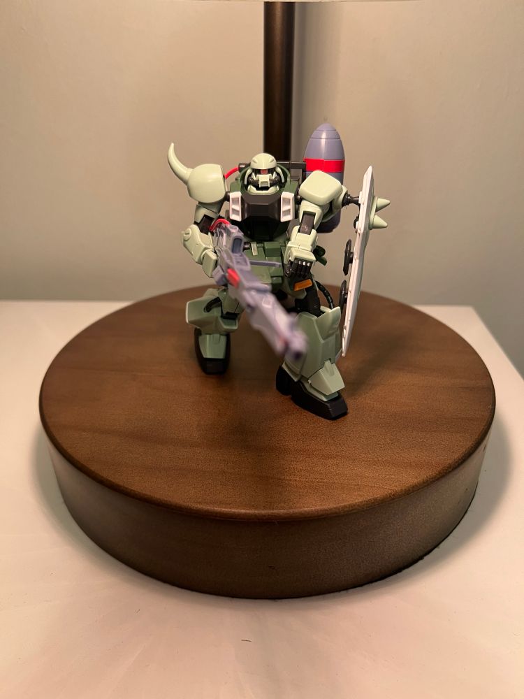 Seafoam green gunner zaku warrior figurine featuring a white spiked shield attached to its left shoulder while holding a long, grey energy rifle with both hands. The weapon is pointed out into the distance.