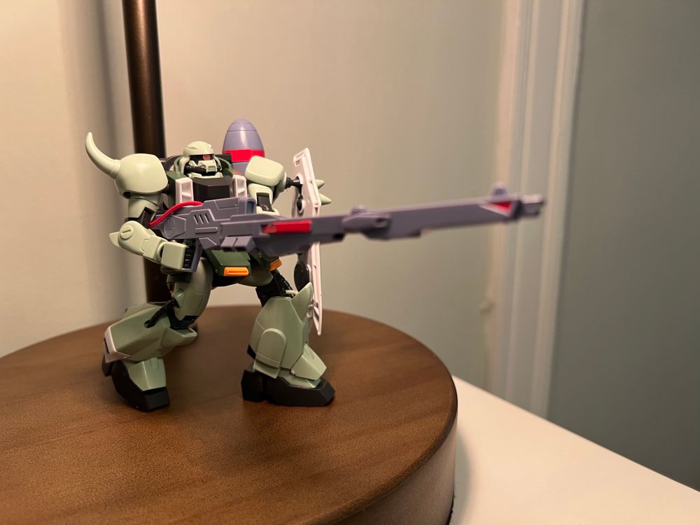 Seafoam green gunner zaku warrior figurine featuring a white spiked shield attached to its left shoulder while holding a long, grey energy rifle with both hands. The weapon is pointed out into the distance.