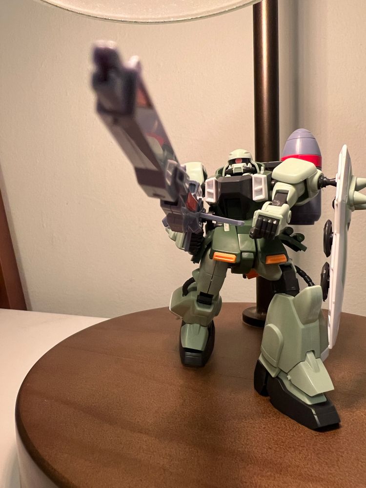 Seafoam green gunner zaku warrior figurine featuring a white spiked shield attached to its left shoulder while holding a long, grey energy rifle with both hands. The weapon is pointed out into the distance.