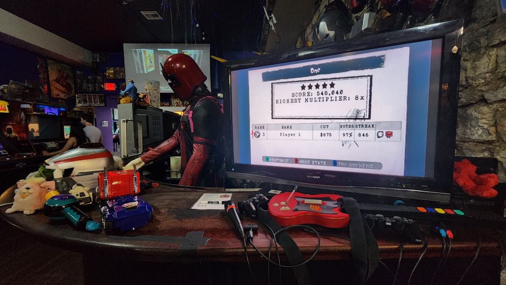 A PlayStation 2 connected to a TV, placed on a bar counter, running Guitar Hero Metallica. In the background are several arcade cabinets and a Deadpool statue behind the TV. 