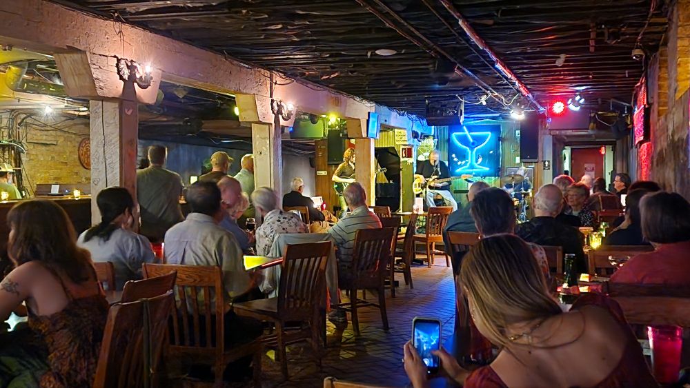 A picture of an underground jazz club. In the foreground are several tables filled with people. In the background is a stage with a guitarist, bassist, and drummer playing jazz (which you can't hear, because this is a picture). Behind the stage is a blue neon sign of a martini glass. 