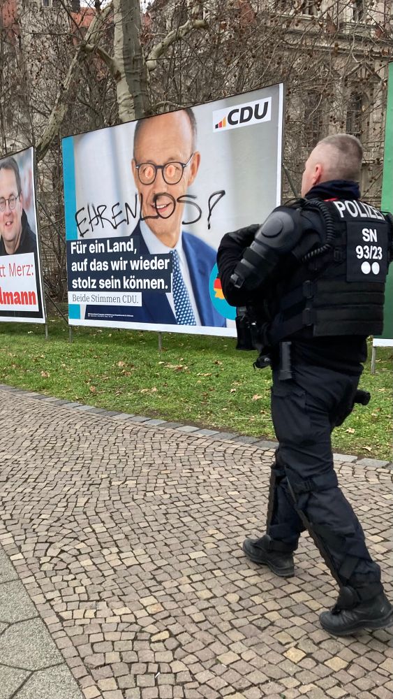 Kreative Verschönerung des Wahlplakats von Merz mit Anus-Äuglein und „Ehrenlos!“-Schriftzug, daneben ein Polizist