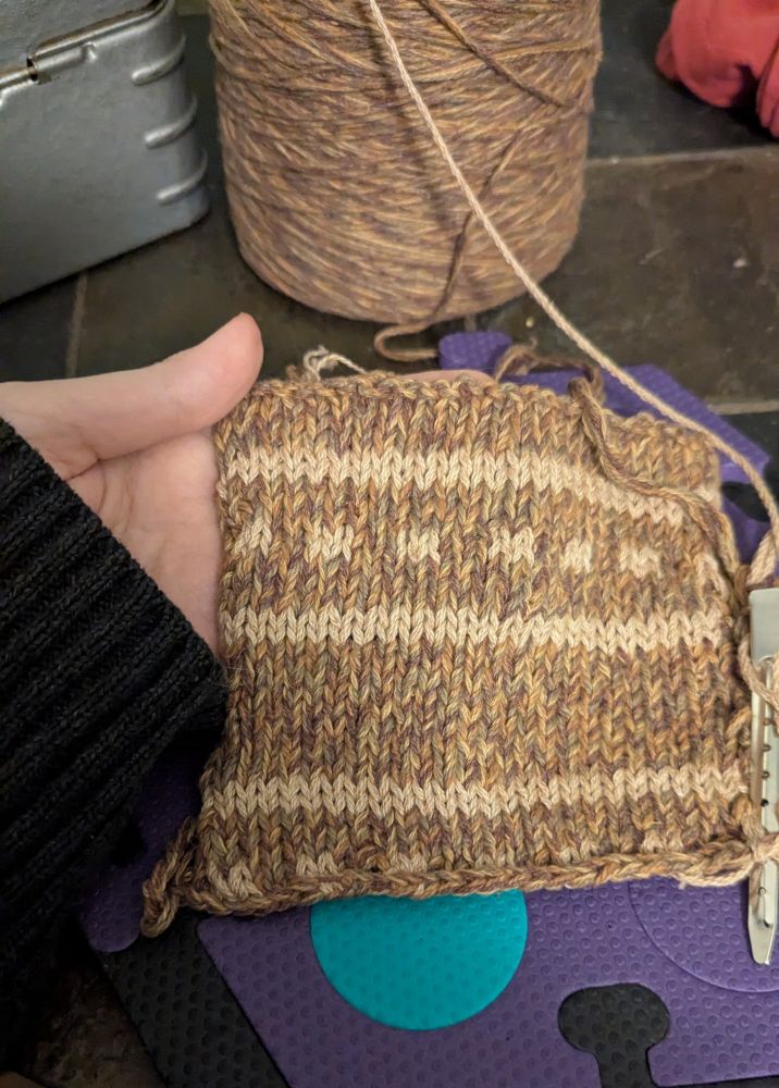 A hand holding a roughly 5"x5" stockinette swatch of cotton. It's a marled brown color with tan stripes and dotted colorwork. The marled brown yarn is four plied with light tan, dark green, reddish brown, and purplish brown cotton strands. 