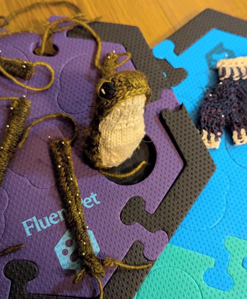 A limbless knit frog sits upright next to a number of knit limbs gruesomely pinned down to foam pieces for blocking. She looks concerned. Part of her sweater can be seen pinned down just out of frame.