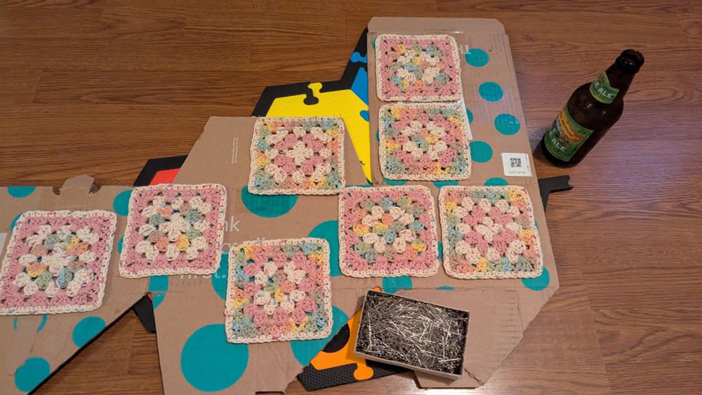 Pink, confetti, and pastel rainbow granny squares pinned to cardboard for blocking on the floor. A beer sits off to the side.