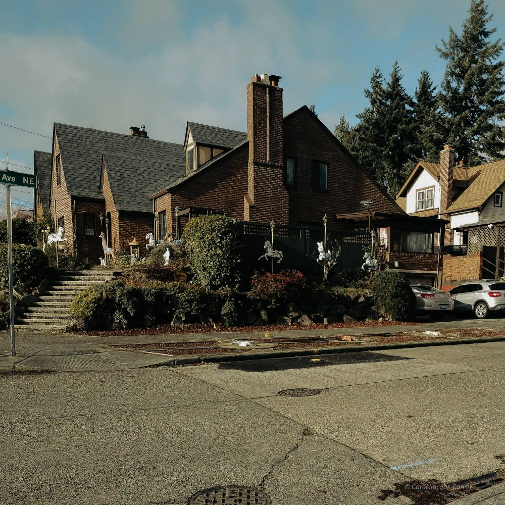 A large brick house takes up most of a corner lot. The owners have decorated the outer perimeter with old carousel animals, including horses, a rabbit and a giraffe, all on their poles. The sidewalk outside the house is quite wide, probably built to spec, with three carousel horses on each of the two street sides embedded in the edging with fancy brickwork framing. I had to stand across the street to get most of the display. The sky above is blue with fluffy clouds. 