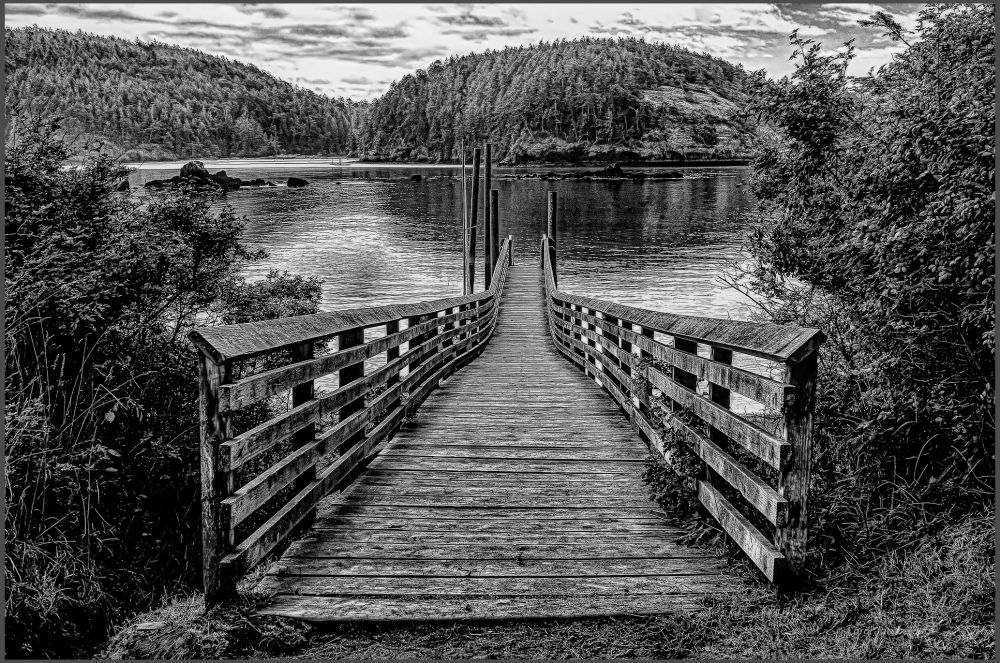 A black and white photo: The boardwalk leads to the ocean inlet at Deception Pass Park in Washington. There are hills covered with pine trees on the other shore, and some bushes at the head of the boardwalk. The water is calm and reflects the sky and hills