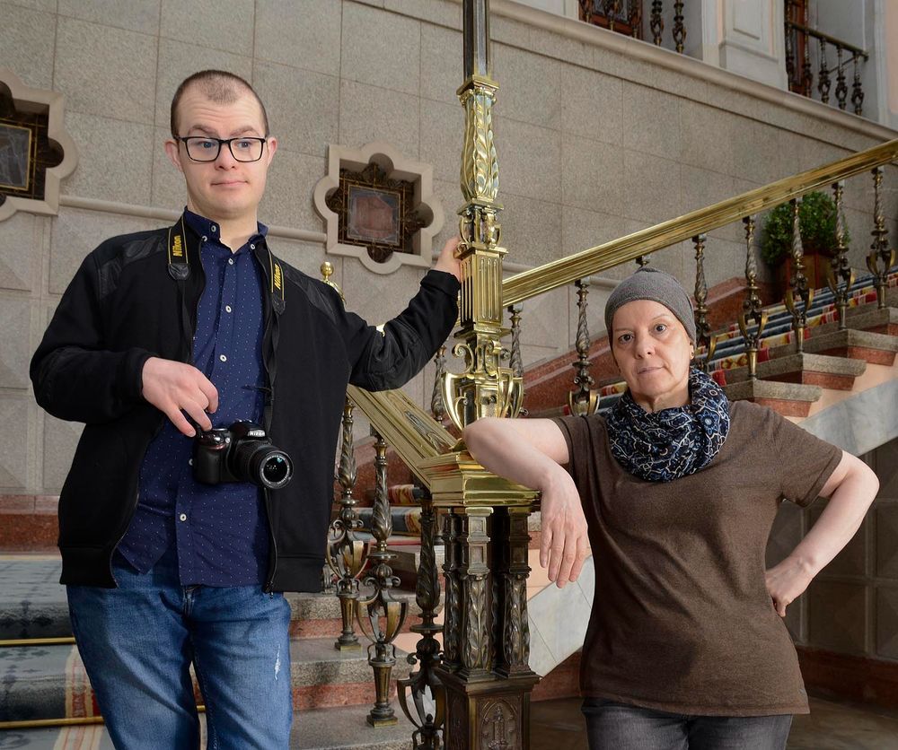 Two people with disabilities, a boy and a woman. The boy has Down syndrome and is carrying a camera. They are standing next to a palace staircase, preparing for a photo shoot. The girl is wearing brown, and the boy is wearing blue.