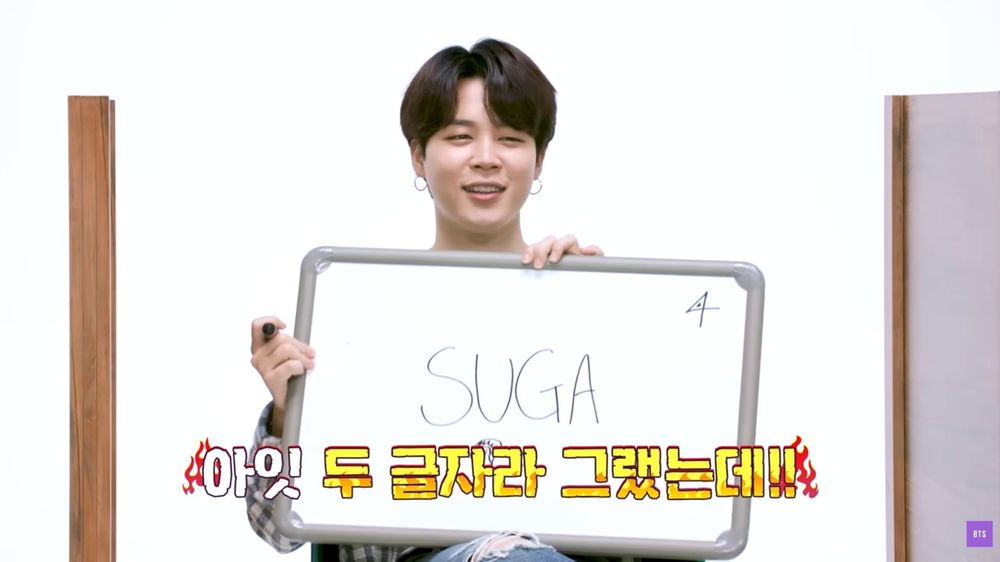Jimin holding a placard with a big writing saying SUGA and a little drawing of a character showing his butt