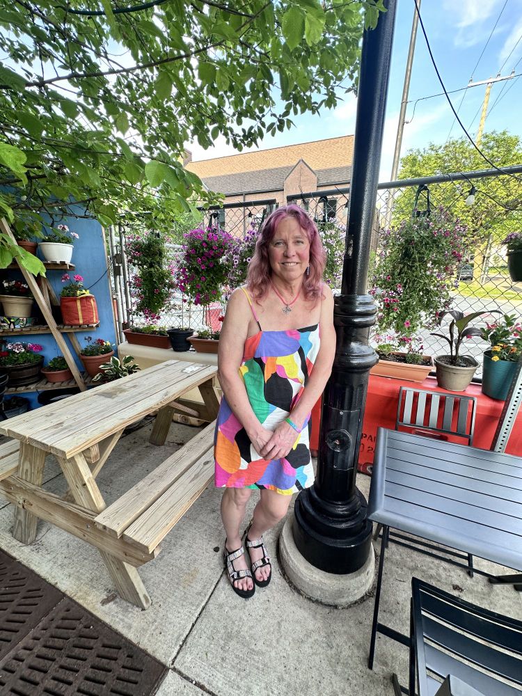 A woman with radiant raspberry hair leaning against a light pole that is black in color. She is outside in a patio area. She is wearing a summer dress with a pattern of solid colors in an array of different shapes. There are green leaves above her. There is a picnic table next to hear. She's wearing multi-colored sandals. It's nice out, so she's wearing a big smile.