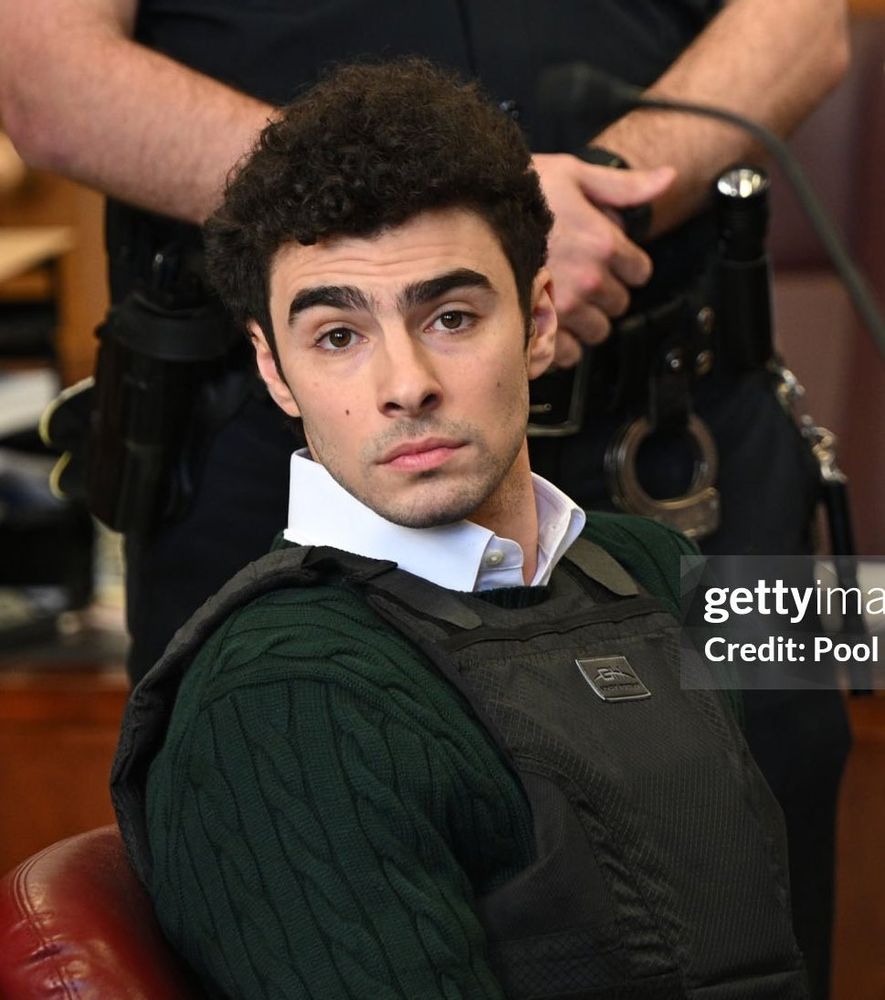 Photo of Luigi Manginess from his hearing today, in a white collared shirt, green sweater and bulletproof vest. He looks directly at the camera in a way that can only be described as "seductive." Behind him stands a guard who is quite likely weak in the knees.