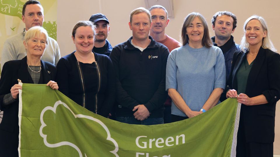 A group of ten people stands together holding a green flag with a tree symbol and the text "Green Flag." They are indoors, smiling, with a mix of casual and formal clothing.