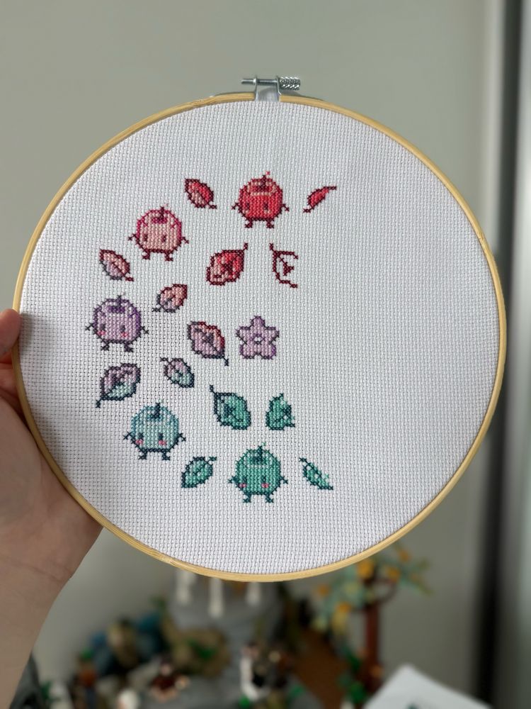 A cross stitch pattern in progress: Stardew Valley junimos and leaves in a pixel art style, circular pattern around a purple stardrop